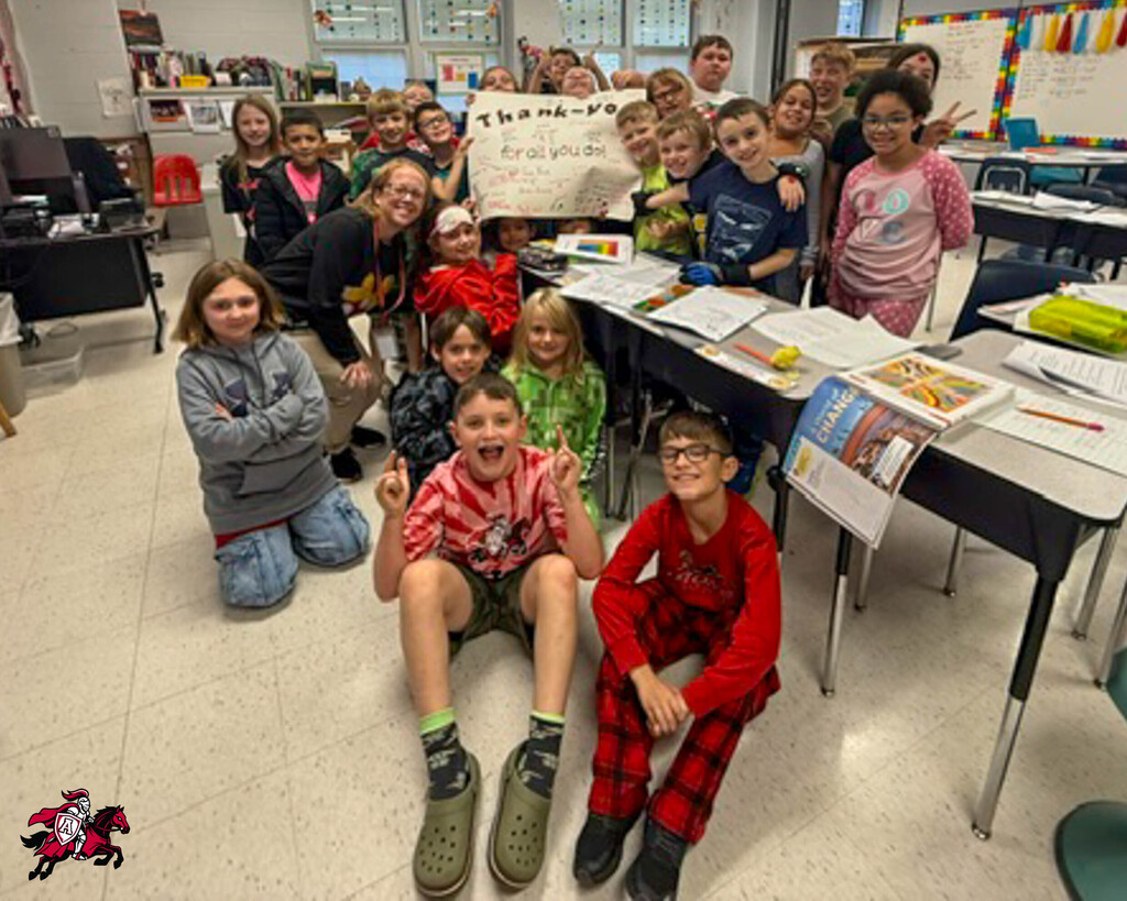 Classroom with students sitting and kneeling. Several hold a white sign. Others stand near tables with papers.