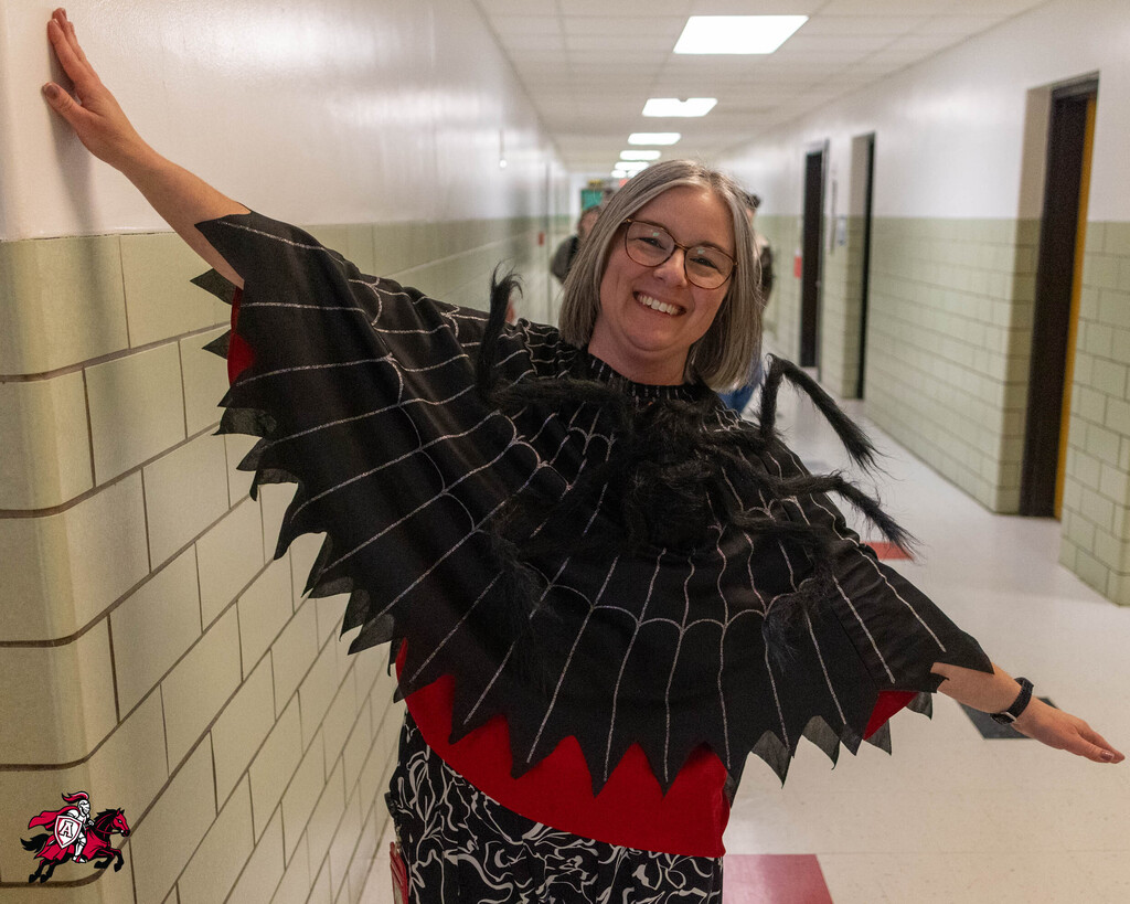 A woman stands in a hallway, leaning against a wall with one arm, wearing a black and red costume.