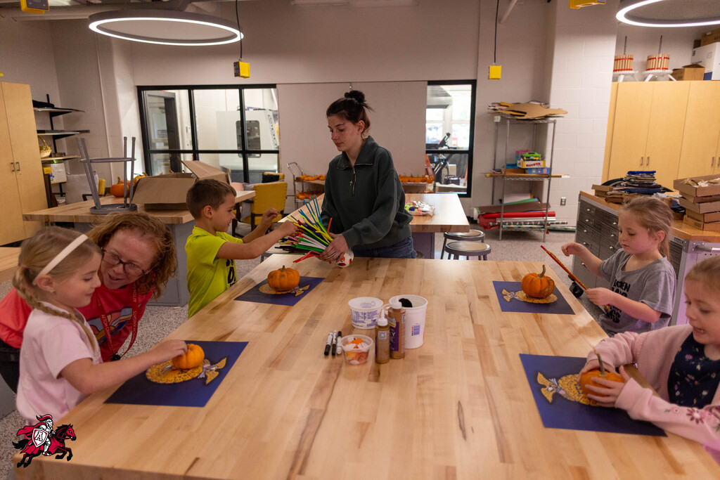 A classroom with children decorating pumpkins. A woman stands at the table helping them.