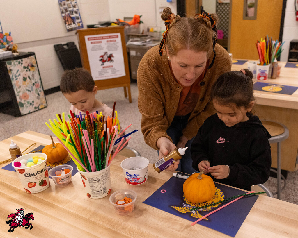 Woman and two children decorating a pumpkin with glue, straws, and small items on a table.