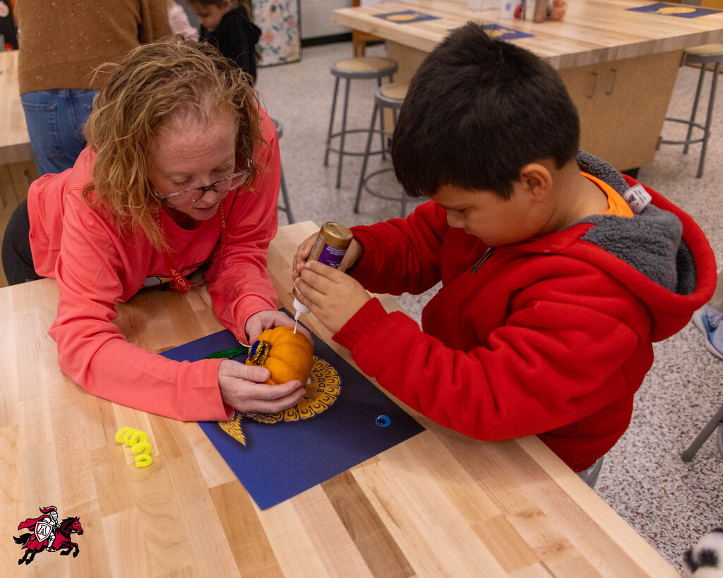 Two children work on pumpkin crafts at a table; one holds an orange pumpkin while the other uses glue.