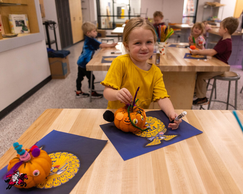 A child seated at a table decorating a pumpkin with a knife and marker.