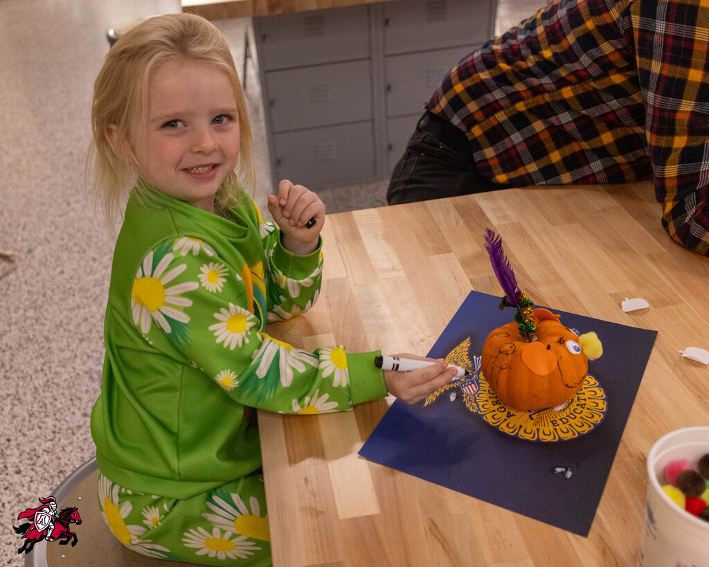 A child in green pajamas sits at a table, decorating a pumpkin with a paintbrush.