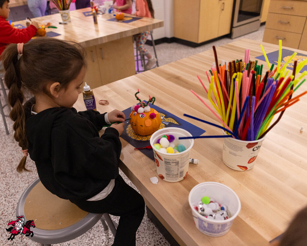 A girl with braided hair uses a glue bottle to decorate a pumpkin with straws and beads.