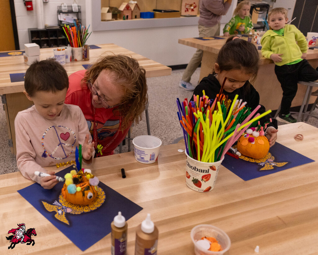 Two children at a table with a pumpkin, glue, and markers. A woman helps one child. Other children work in background.