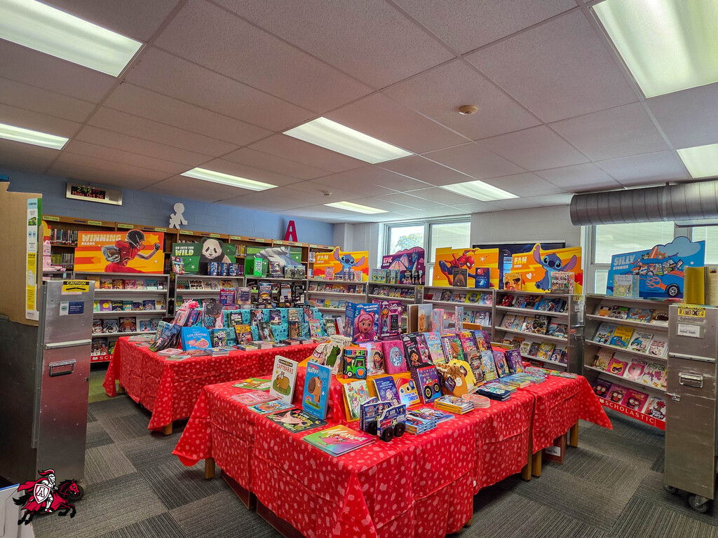 Library with multiple tables covered in red fabric, displaying a variety of colorful books and toys.