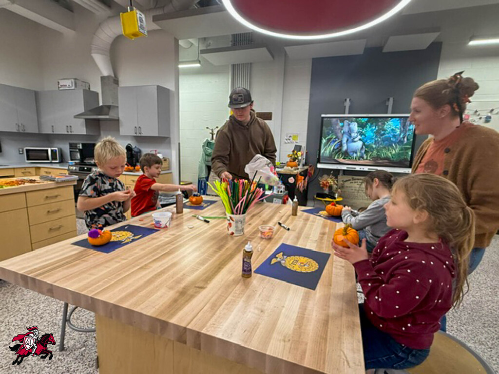 Adults and children sit around a table with pumpkins, paintbrushes, and other craft supplies.