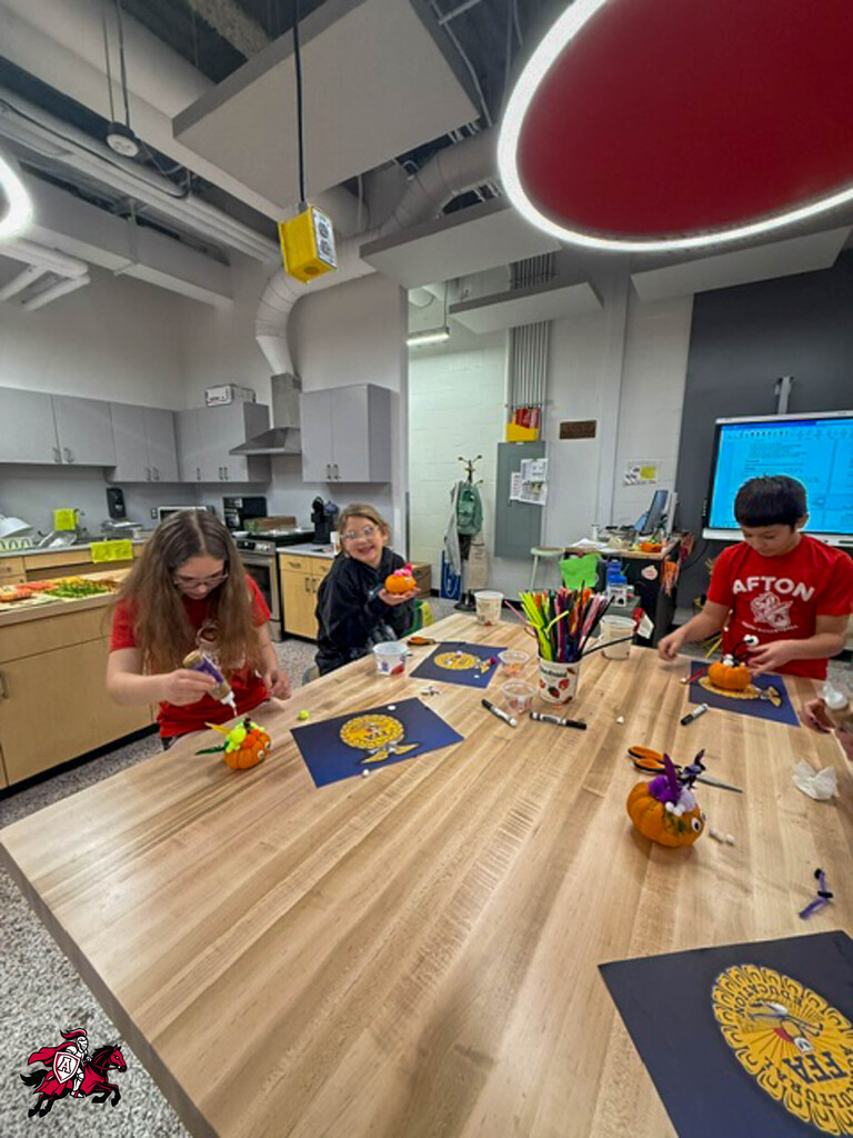 Three students around a table with pumpkins, paintbrushes, and paper. Bright room with wooden table and cabinets in the background.