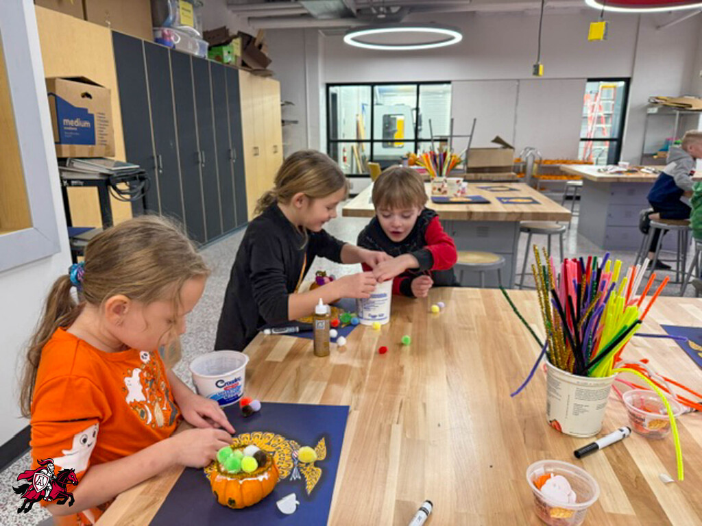 Three children around a table; one with a pumpkin, one with glue, and one with colorful plastic tubes.