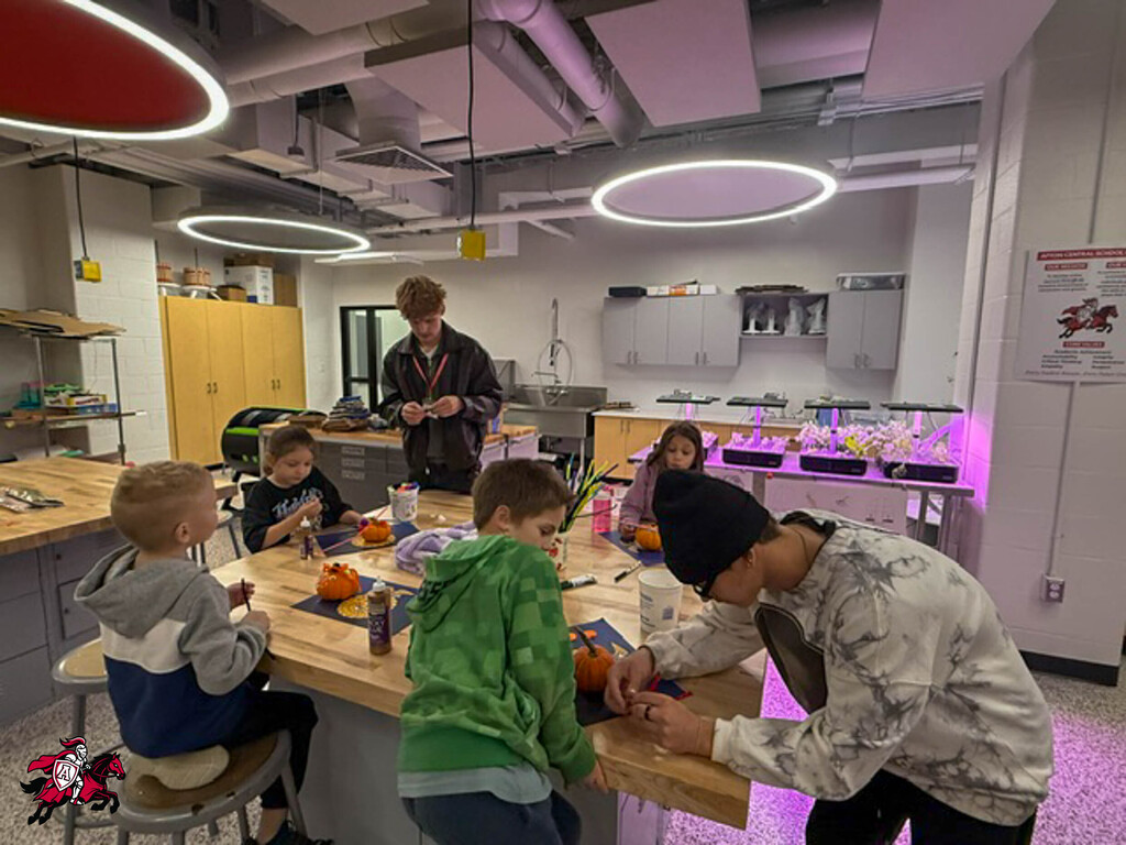 Several children sit around a table with pumpkins while an adult holds a knife. The room has overhead lighting and a plant in the background.