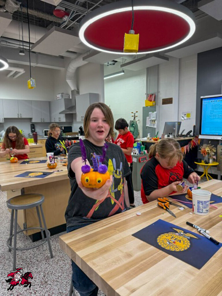 Several students work on crafting pumpkins in a classroom. One student holds a pumpkin with eyes and a nose.