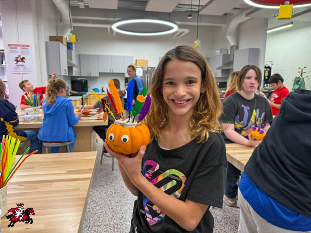 Girl holding pumpkin with feathers, smiling. Room with tables, chairs, and other people. Colorful cups and straws on table.