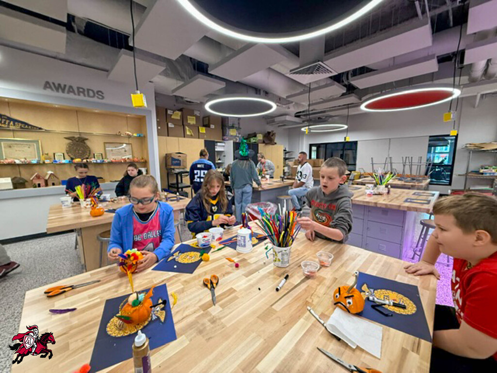 Students work at tables, carving pumpkins with utensils. A wall behind them displays awards.