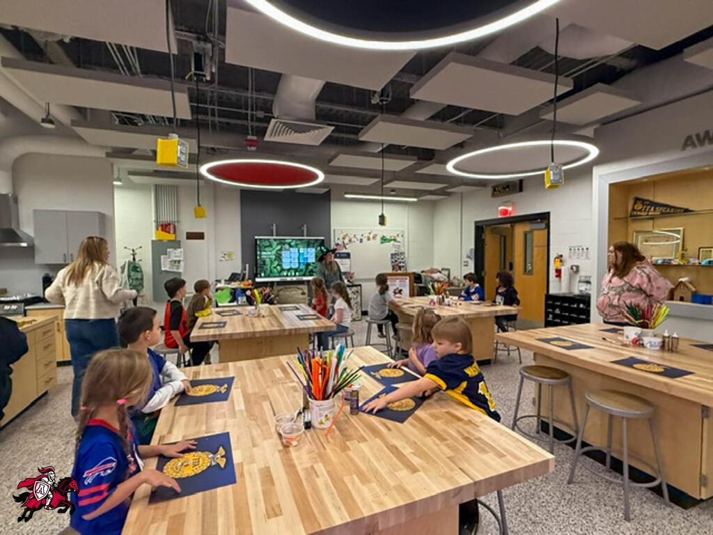 Children sitting at tables in a classroom with pencils, markers, and paper. Adult standing nearby.