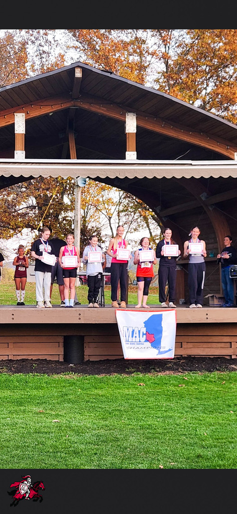 A group of young athletes holding signs stand on a stage under a wooden roof. A banner hangs in the foreground.