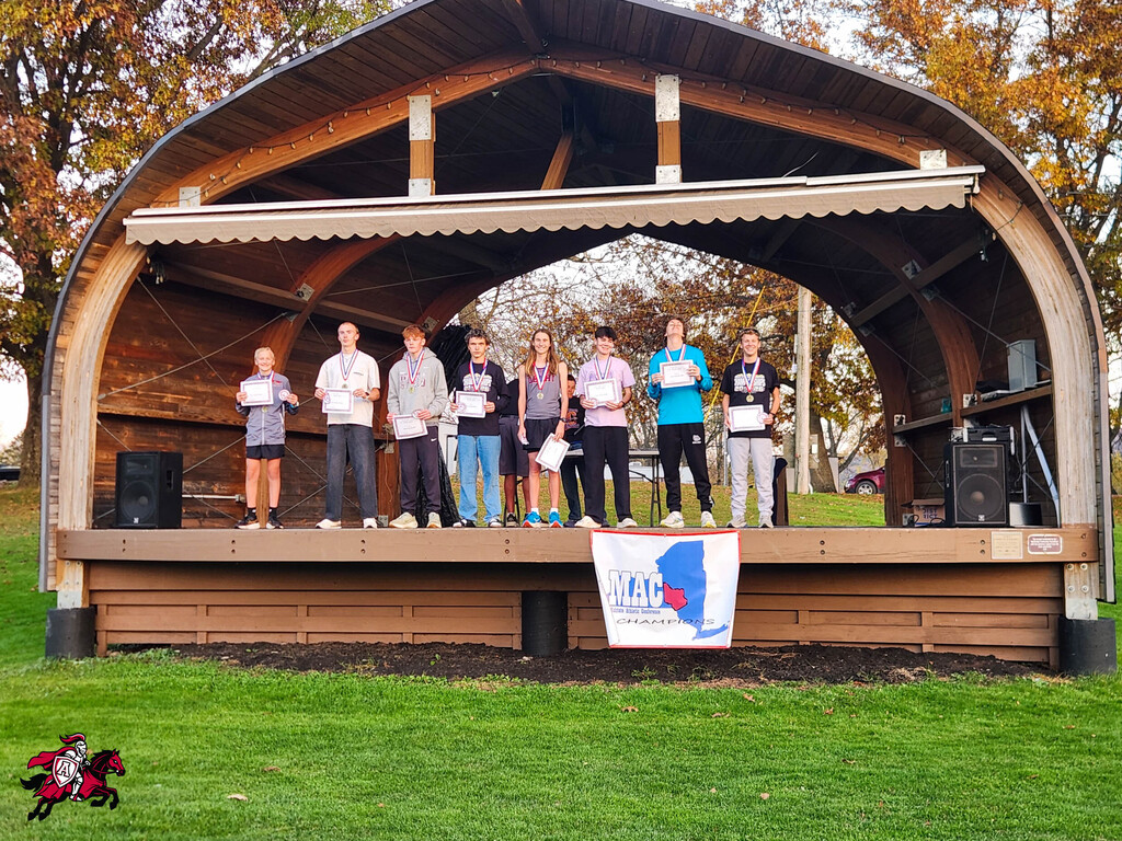 People stand on a wooden stage holding certificates. A banner is displayed in front.