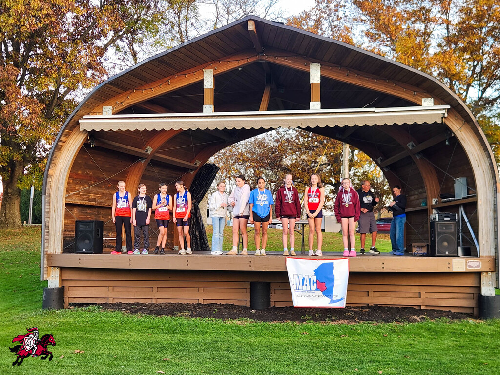 People stand on a stage with a banner, speakers, and a roof; some wear athletic uniforms.