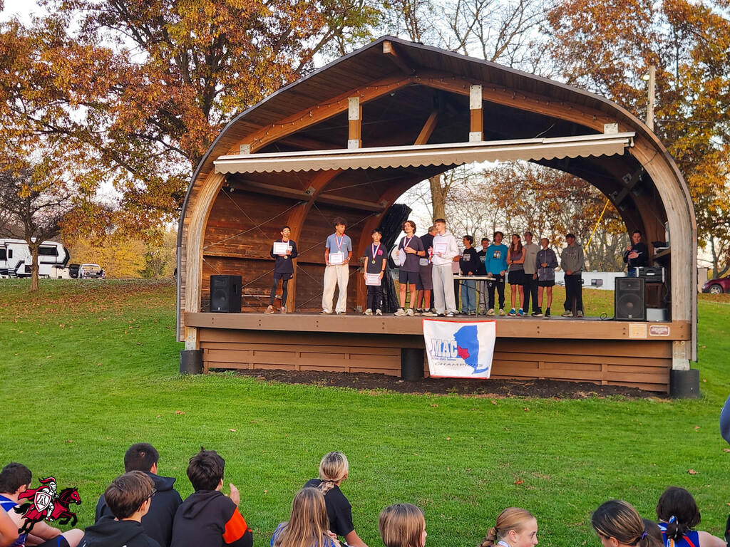 Students gather on a grassy area; some stand on a wooden stage with a flag in front.