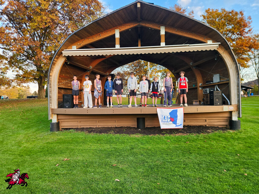 Nine children on stage, standing in front of a wooden arch with a banner, on a grassy field.