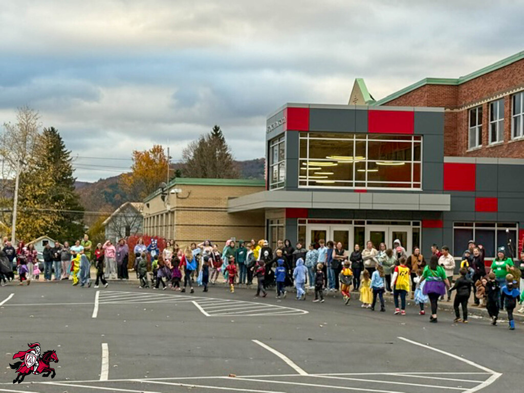 Many people wearing costumes and carrying bags walk in a school parking lot with trees in the background.
