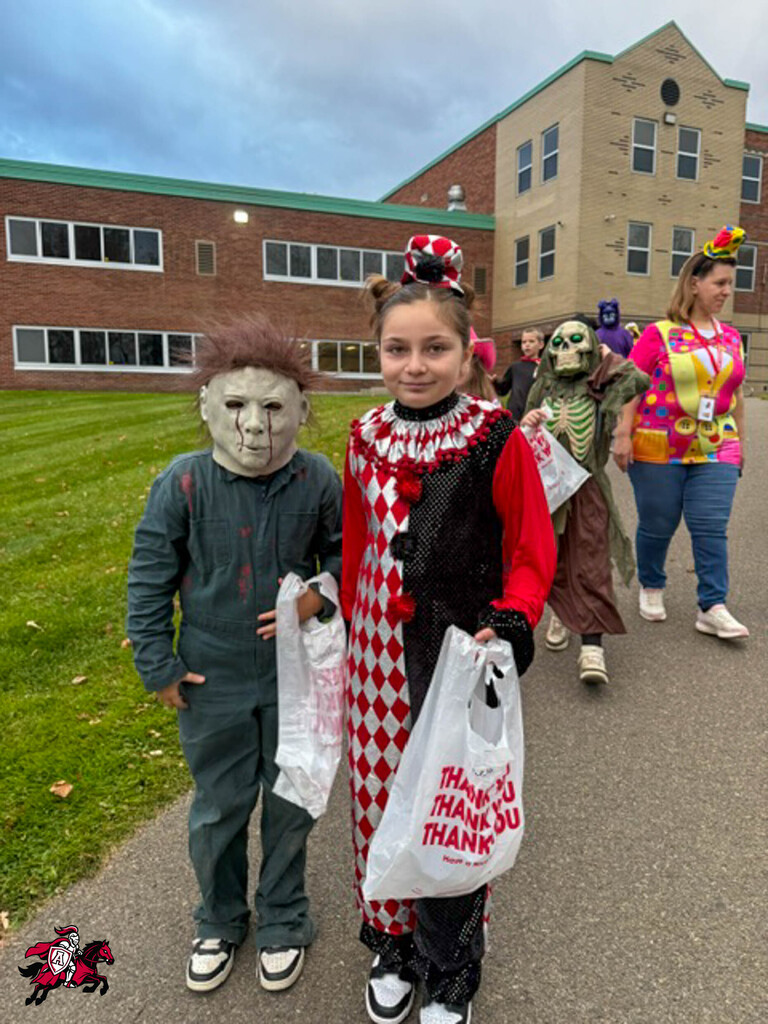 Children in costumes walk past a school building with large windows, green grass, and a cloudy sky.