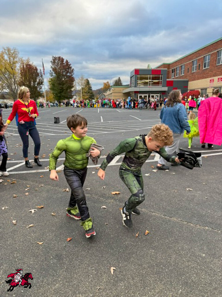 Two children in superhero costumes run in a parking lot. A person in red and black holds a baseball glove.