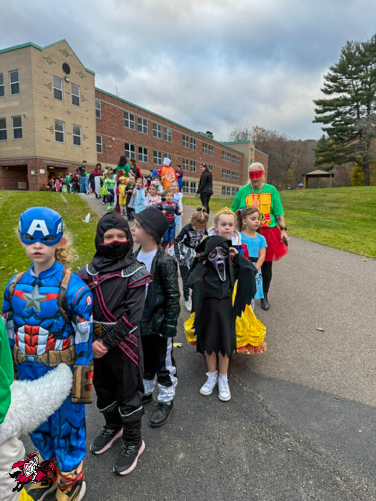 Children in costumes walk on a sidewalk in front of a building, with a grassy area behind.
