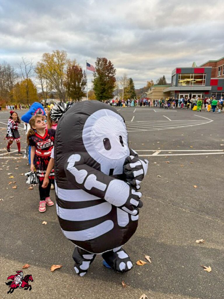 Person in a skeleton costume stands in a school parking lot. Two children in costumes walk behind them.