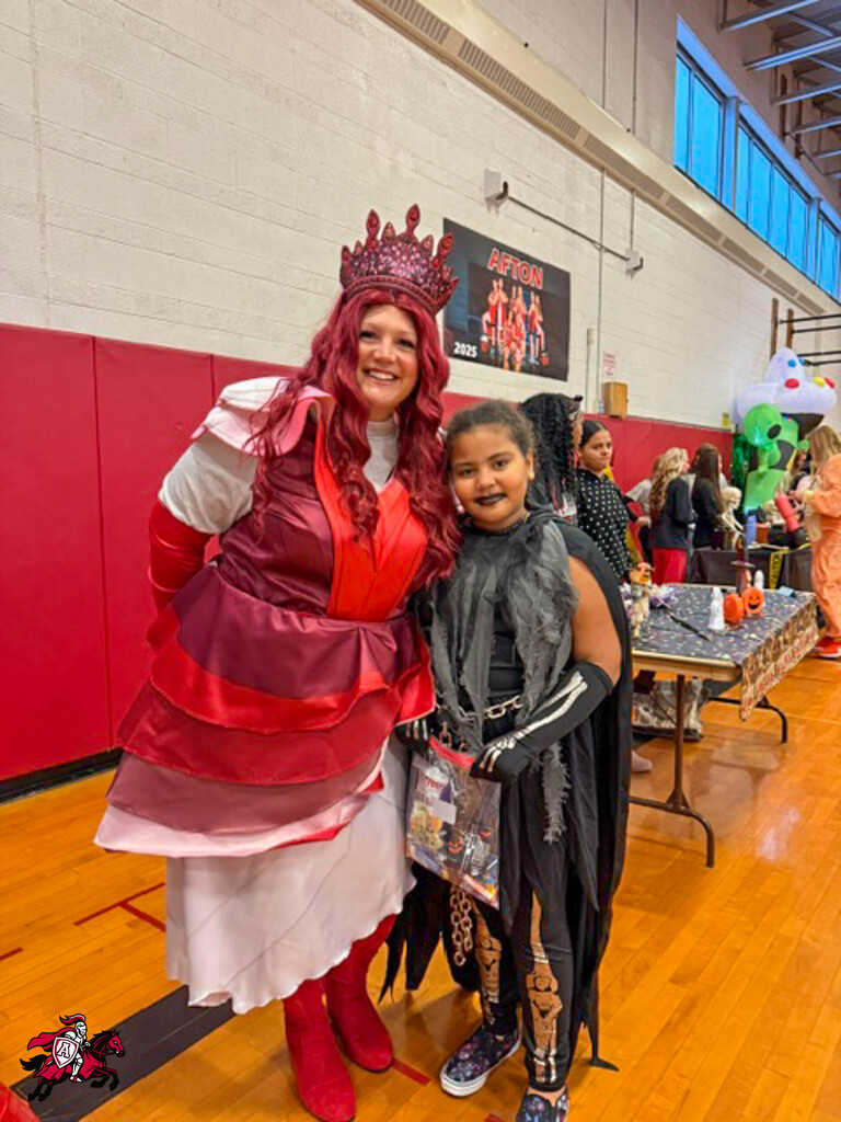 Two people in costume in a school gym. One wears a red gown, crown, and boots. The other wears a black costume.