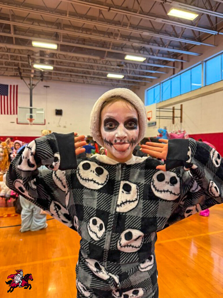 A person with face paint stands in a gym wearing a Jack Skellington costume, holding hands to shoulders.
