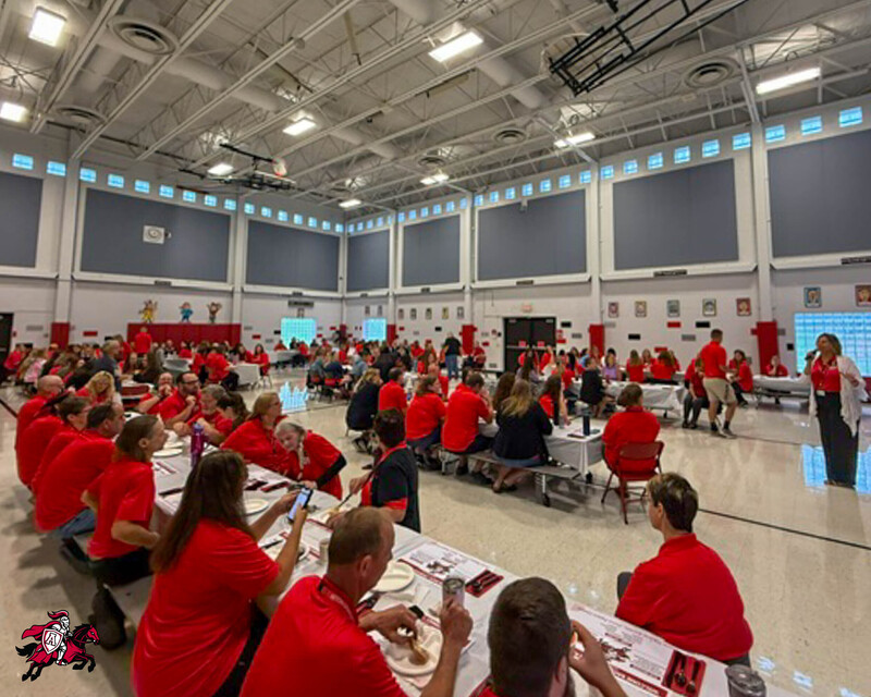 All Afton Central School District teachers, faculty and staff gather in a cafeteria for breakfast as Superintendent Annette Hammond speaks into a microphone at the front of the room