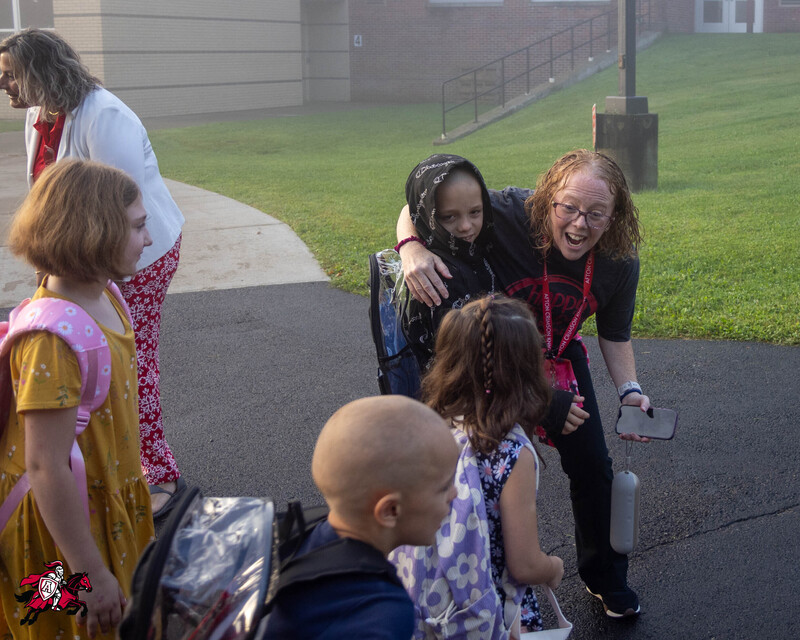 A group of students are greeted by a principal as they step off a school bus on their way into a school building, as the principal puts her arm around one of the students in the group