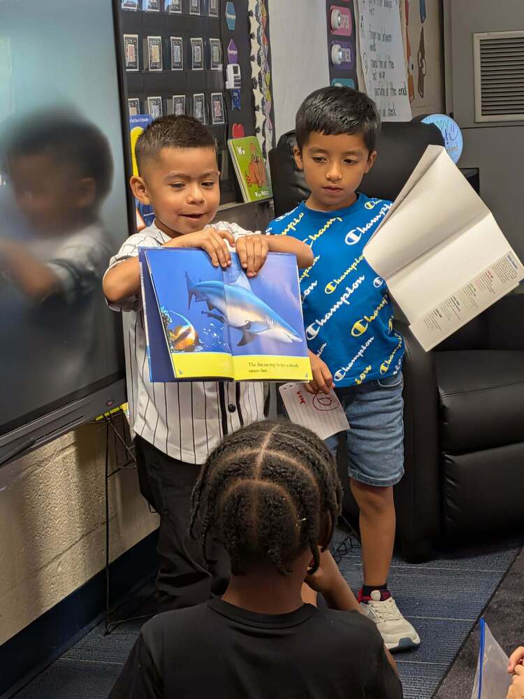 Two boys are holding up a book in front of their class.