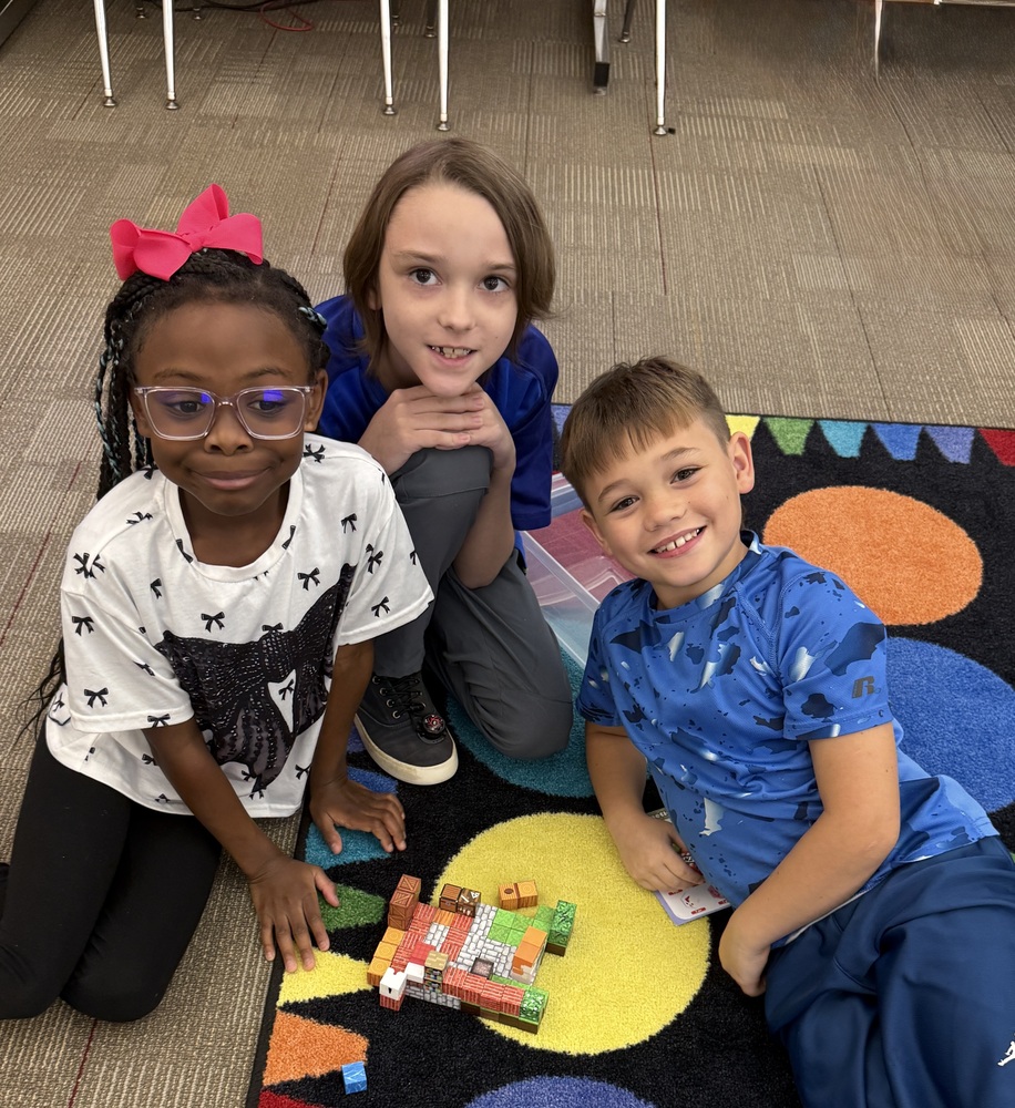 Two boys and a girl are smiling and sitting on the floor next to some blocks.