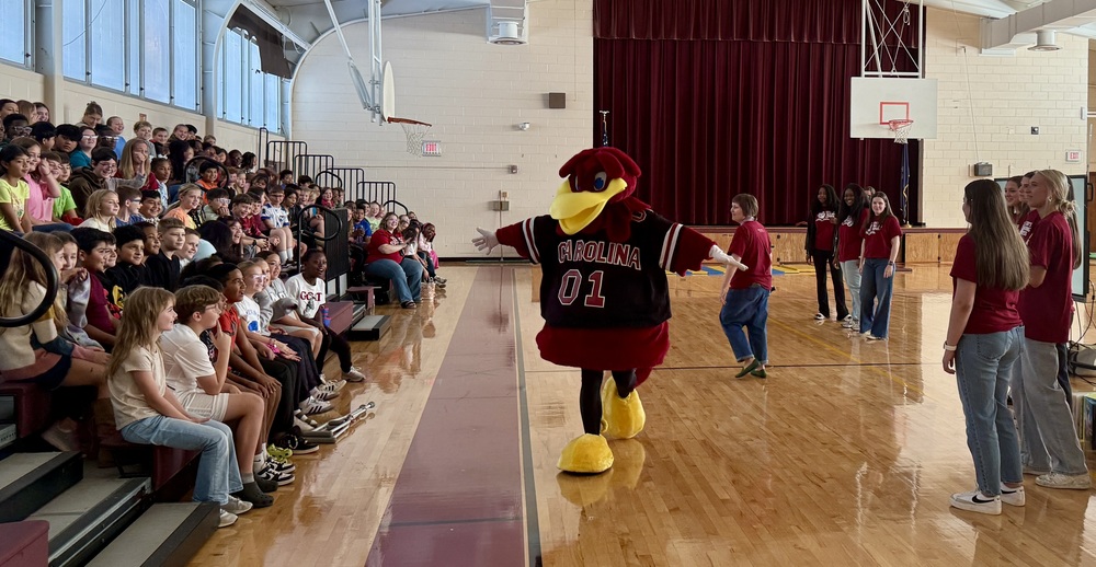 Students seated on bleachers watch a presentation by Cocky's Reading Express with college students and the mascot, Cocky.