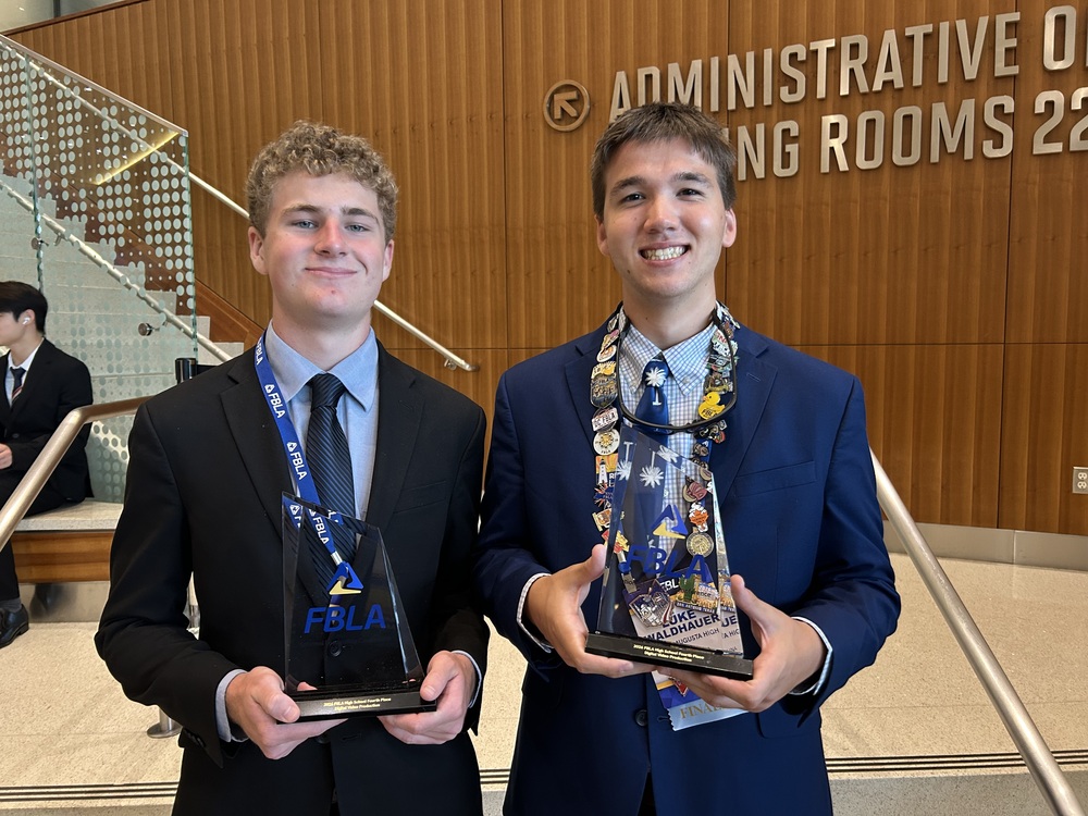 "Jacob Seigler and Luke Waldhauer in suits in front of stairs holding award."