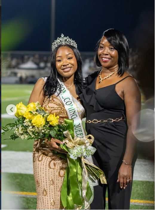 Girl holding flowers and wearing a crown smiling and standing next to a lady in a black dress. 