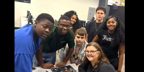 Students surrounding teacher working on a laptop in classroom. 