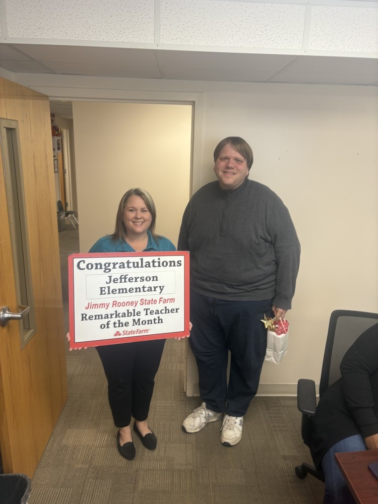 A woman from Jimmy Rooney State Farm holds a sign for Teacher of the Month next to a male teacher.