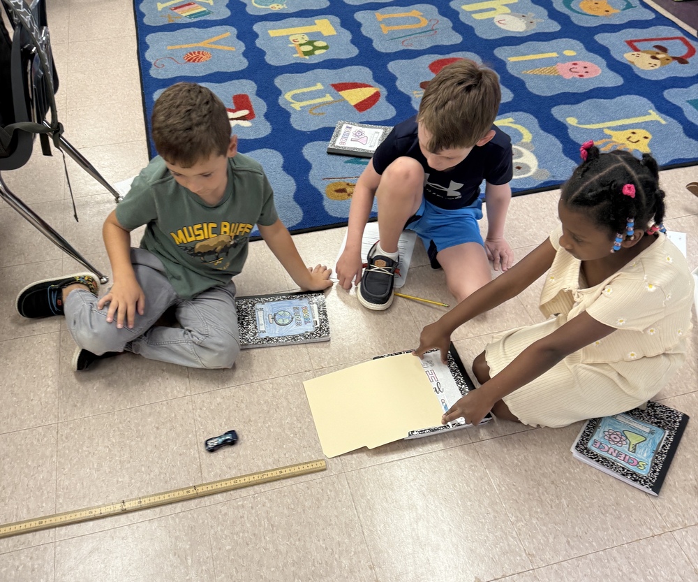 Three students sit on the floor conducting a science expperiement with a toy car.