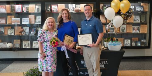Counselor surrounded by principal and announcer in front of display case with table and balloons. 