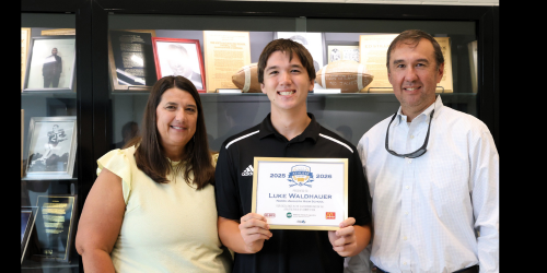Mother and Father on side of student holding award in front of trophy case.