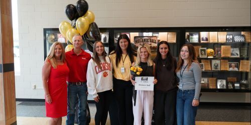 Parents and a group of 5 girls in front of trophy case with black and gold balloons. 1 Girls is holding a paper award.