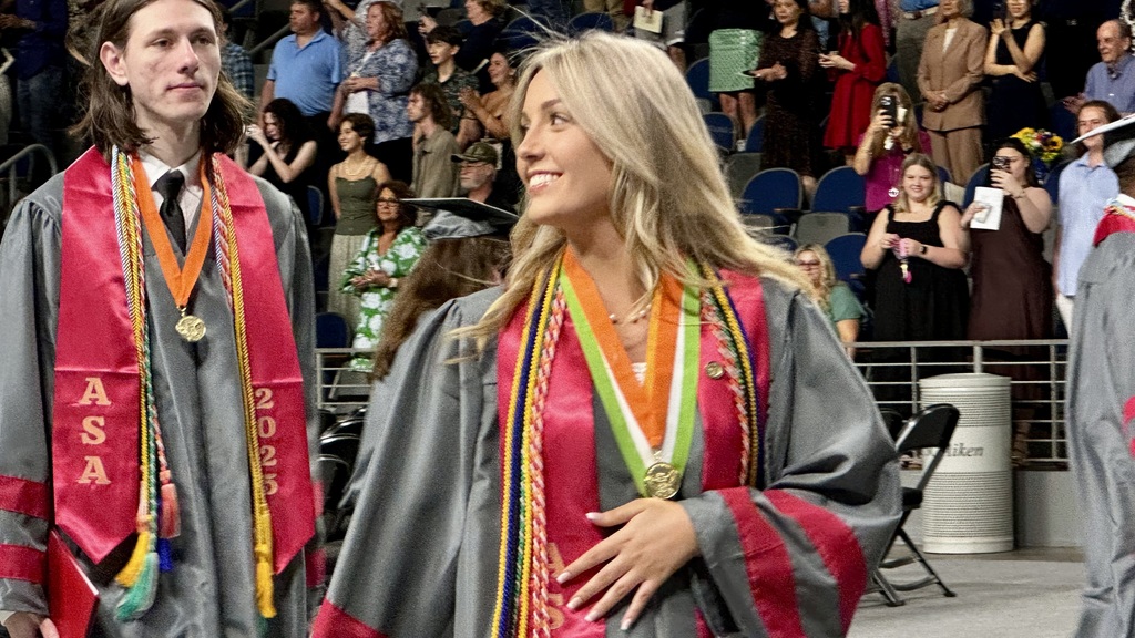 Blonde female graduate wearing a gray robe with burgundy accents looking into a crowd.