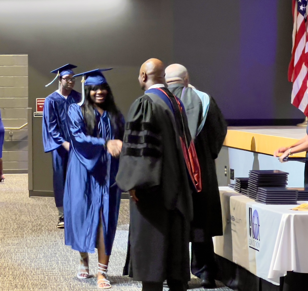 Female Adult Graduate in a Blue Robe and Cap shaking hands with the Superintendent in a Black Robe.
