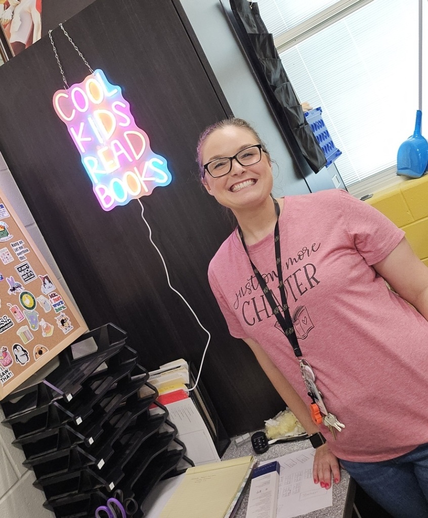 Ms. Odenthal standing behind her desk with neon sign reading cool kids read books. 