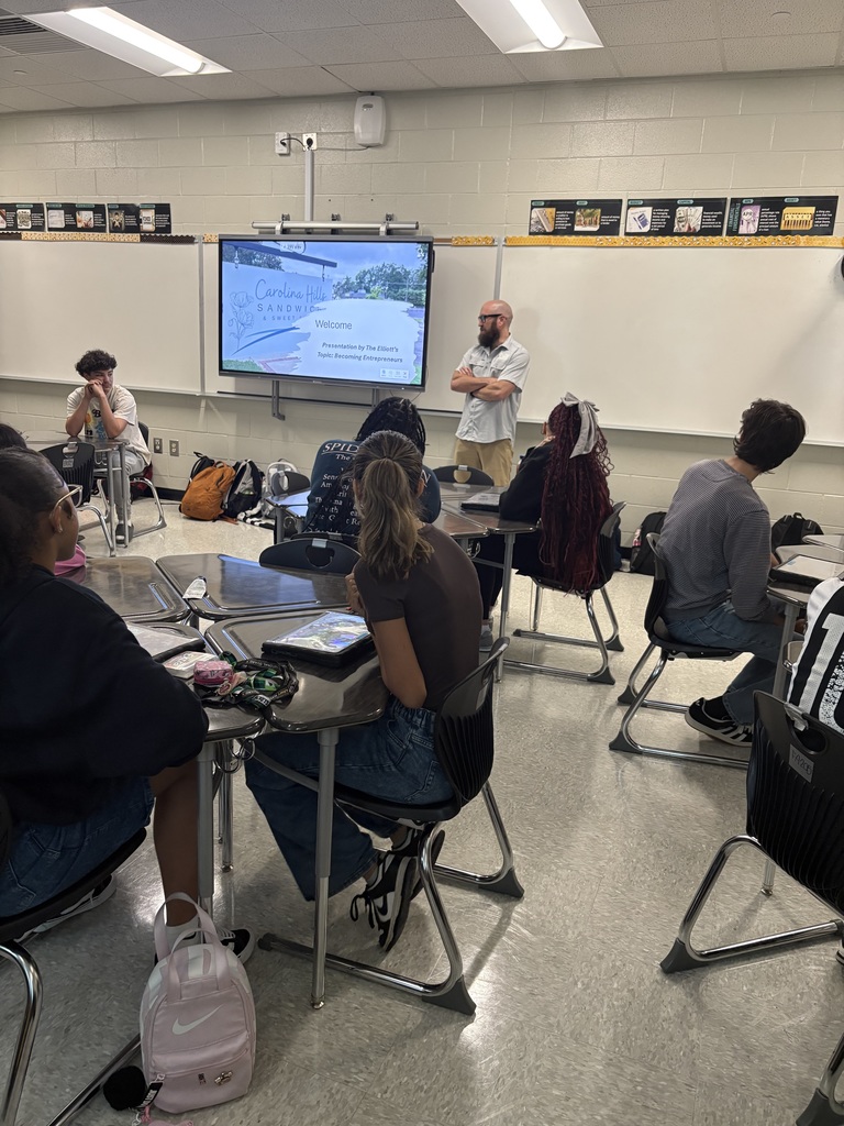 Man with beard speaking to a group of students sitting at desks. 