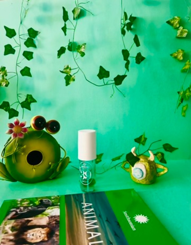 Green based photo with metal abstract frog, perfume, horned clock, and animal book with green ivy. 