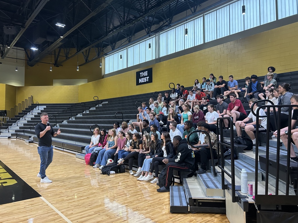 Group of students sitting in gym listening to male speaker. 