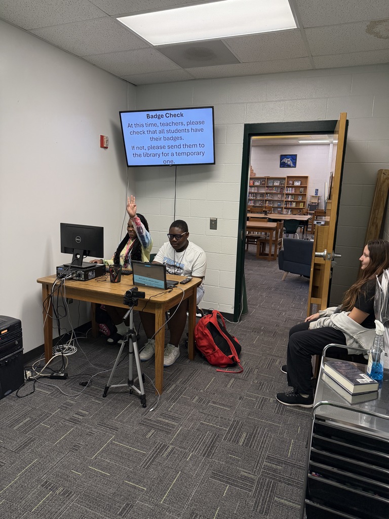 one student holding up her hand in a count and another student working on a computer. They are leading a news crew. 